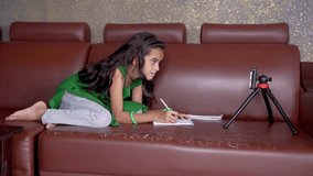 Concept of improper, lazy and way of studying during homeschooling or e-learning and make kids sleepy or boredom, young girl busy in writing by looking into mobile on sofa during covid-19 pandemic