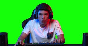 Excited professional young asian male cyber sport gamer in headset with a mic concentrated playing in online video game in colourful UV lighting over green screen background. 