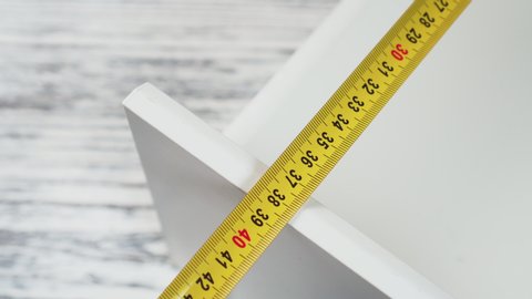 Measurement of furniture with a yellow metal measuring tape. White cabinet shelf on wooden background close-up