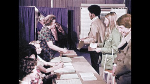 1970s: People wait in line at table to vote while people vote in booths behind them. Person goes into voting booth. Person comes out of booth. Instructions on wall to vote. Proper, improper marks.