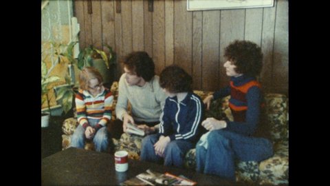 1970s: Man sits on couch with two children, reads story to children. Woman wipes down coffee table, sits on couch. Woman talks and smiles.
