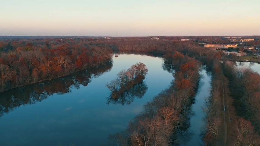 Drone circling island in still Lake Carnegie surrounded by a US northeast forest of fall, orange colored trees in Princeton, New Jersey. Taken at sunset during autumn.