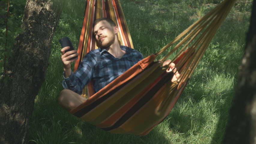 Mobile internet surfing. Man with cell phone in hands relaxing in hammock outdoors. Relax in hammock. Young male tourist lying on hammock and using mobile phone. Camping. Summer vacation