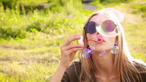 Close-up portrait of an attractive elegant caucasian blonde in sunglasses lazily and slowly blows soap bubbles, sits on the lawn at the public park, looking at camera, relaxing after hard working day.
