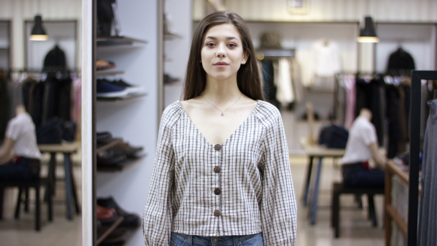 A front view close up shot of a young beautiful woman standing in a clothing boutique lookin straight into camera and changes her pose several times
