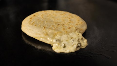 Camera pans back in slow motion to reveal a hot Salvadoran pupusa cooking on a griddle. The cheese filling oozes out, spilling over the hot surface with a sizzle.