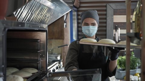 small business, young cute female baker in medical mask puts bake with slices of dough for baking in an electric furnace while working in bakery after removing quarantine