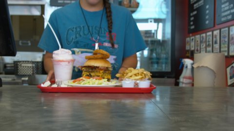 Woman employee at a burger joint pushes a full tray with the largest burger and fries and shake across the counter for the customer to pick up.