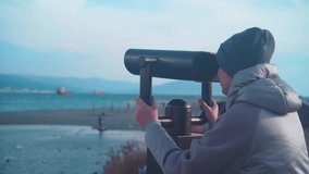 A young woman in a hat looks at the sea, mountains and ships in a binocular, atmospheric video. Sightseeing.