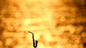 Silhouette of an alto, tenor saxophone stands against backdrop of golden sunset or dawn. Beautiful golden blurred background, game of sun glare of sun on sea water. Musical theme. Slow motion video.