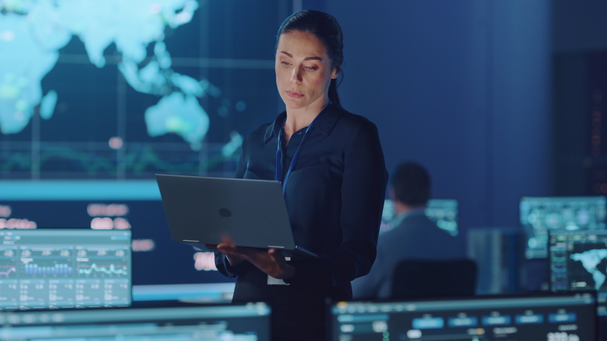 Female Project Leader is Standing with Laptop Computer and Checking Work Data. Science Engineers Work Around Her. Telecommunications Control Monitoring Room with Neural Network on Servers. | Shutterstock HD Video #1055506658