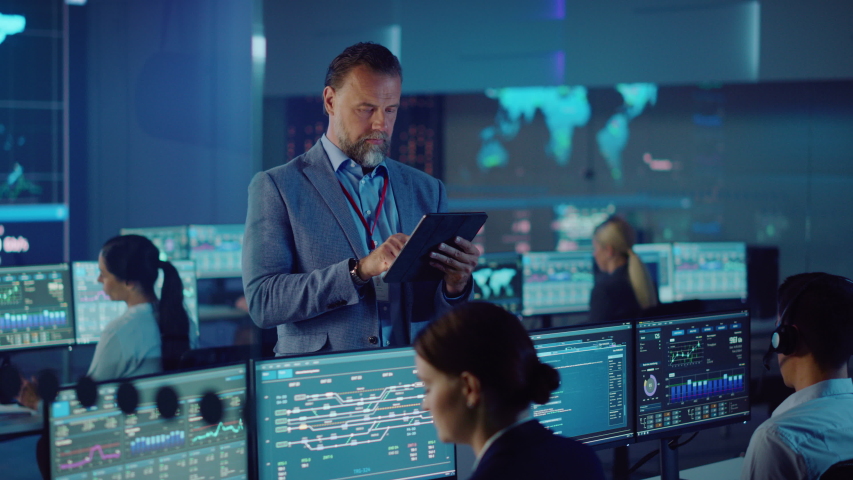 Male Project Leader is Standing with Tablet Computer and Making Mental Calculations. Data Science Engineers Work Around Him. Telecommunications Control Monitoring Room with a Global Map on Big Screen. | Shutterstock HD Video #1055506853