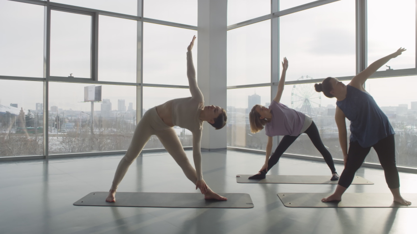 Group of three modern young women with differnet levels of body flexibility practising yoga in spacious hall with panoramic windows, back light shot | Shutterstock HD Video #1055510999