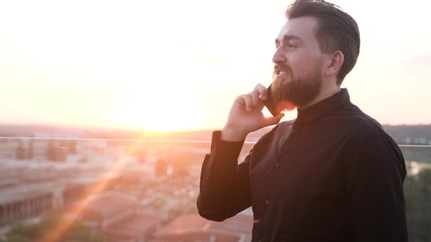 Businessman staying on his house roof and talking on phone with his partner, admiring the view, sunrise/sunset and city view from the high