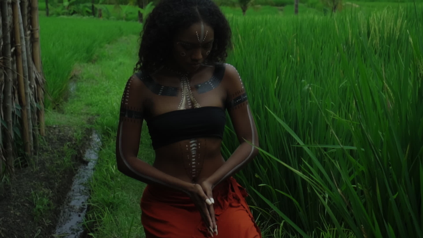 Video of a beautiful young brunette Africa girl with makeup and painted etрnic lines on the body walking in a tropical garden