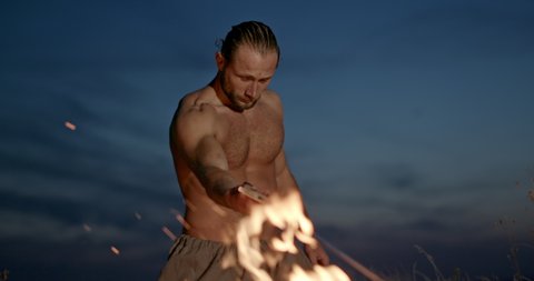 Barbarian warrior heating sword in fire. Tilt up view of strong shirtless man heating sword in burning fire then lifting blade to face against cloudy evening sky in wilderness