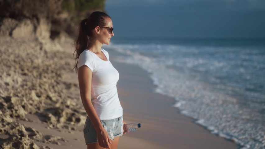 Beautiful young woman in sunglasses drinks clear water from plastic bottle on ocean beach. Blured waves and rock on background. Girl quenches thirst on hot summer day Royalty-Free Stock Footage #1055532152