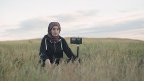 A Muslim girl blogger with a headscarf takes pictures of herself on a phone on a tripod and gives a thumbs up. Young girl Muslim blogger shoots video in a field in the fresh air