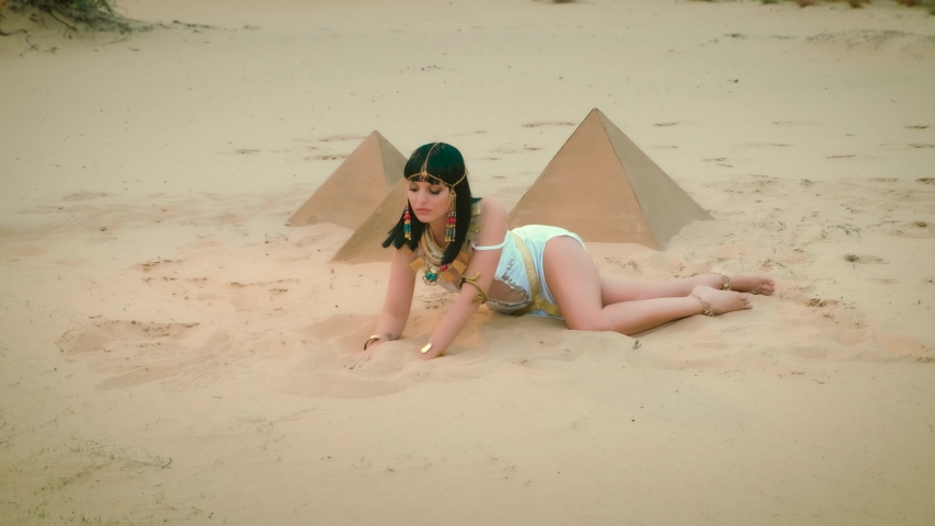 Beautiful young woman fashion model posing, image pharaoh Queen Cleopatra lies on sand desert. Sexy bare legs White antique dress costume, gold jewelry tiara necklace. black loose hair bob haircut