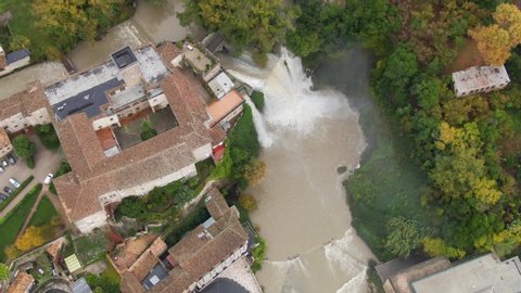 Overhead view of Isola del Liri waterfall cascading down into brown pool of water with view of downtown Lazio, Italy, above rising aerial tilt up