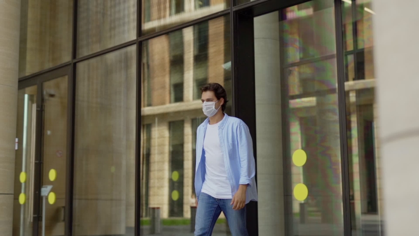Panning slow motion shot of young man walking out of building and taking off medical face mask outside