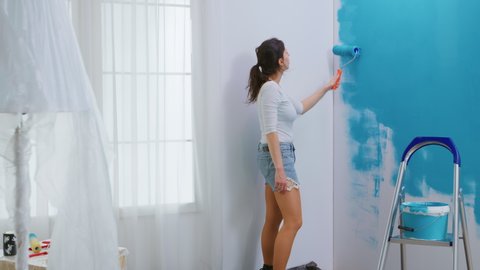 Adult woman painting wall using roller brush dipped in blue paint. Home designer renovate, renovating. Apartment redecoration and home construction while renovating and improving. Repair and