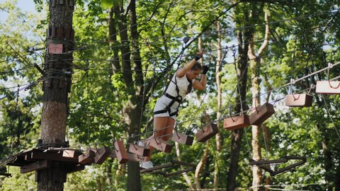 Brave teenage girl overcomes difficult obstacles while walking along a rope stretched between tall trees in a rope town