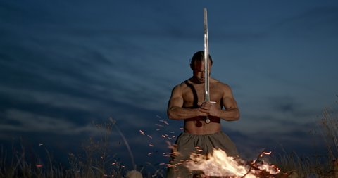Barbarian warrior with sword near campfire. Muscular shirtless man with sword kneeling near burning fire against cloudy evening sky in nature