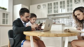 Comfortable Life of Family Together Sitting at Table in Kitchen. Serious Business Man Looks at Phone Discussing Remote Works on Video Call Using Laptop During Holding Little Son While Wife Reads Book