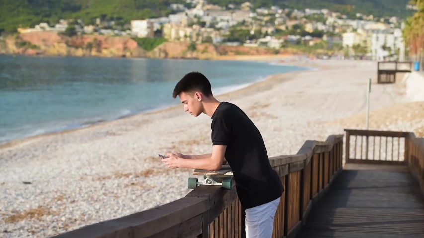 Teenager with a smartphone and a longboard on a beautiful wooden walkway at dawn, giving access to a round stone beach on the Mediterranean coast
