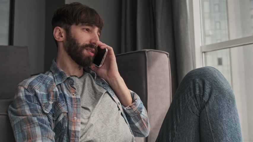 A positive cheerful young hipster man in casual clothes is talking on his smartphone sitting in the living room at home | Shutterstock HD Video #1055567423