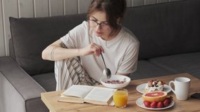 A beautiful woman wearing glasses and pajama is reading book during breakfast in a cozy room at home in the morning