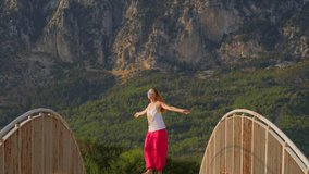 Young slender girl in bright pink Indian wide trousers, white T-shirt, with long blond hair, is dancing, spinning on bridge, amid mountains in early morning. Summer vacation, fun. Slow motion video.