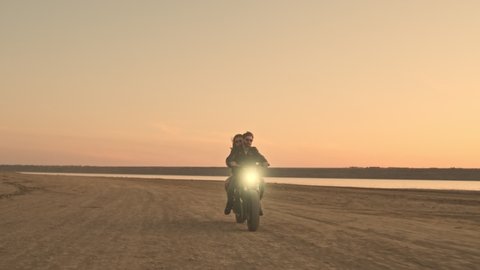 A stylish young couple man and woman are riding on a motorbike together on the beach during sunset time with headlight on