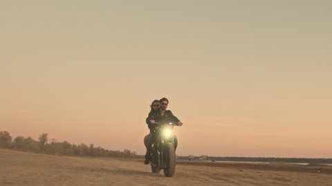 A happy young couple man and woman are riding on a motorbike together on the beach during sunset time with headlight on