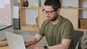 A calm handsome young businessman wearing glasses is working with his laptop sitting at the table in the office