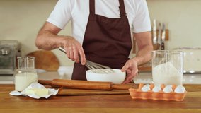 A good-looking mature man in apron is whisking dough at a cozy kitchen