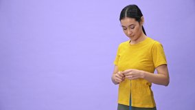 Happy pretty sportswoman in yellow t-shirt holding measuring tape on her waist and rejoicing after that over violet background