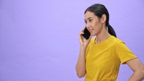 Cheerful pretty woman in yellow t-shirt talking by smartphone and looking away over violet background