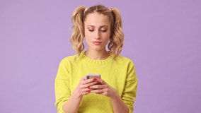 A lovely blonde girl is typing on the phone and rejoices at the answer of the interlocutor in an isolated studio on a purple background