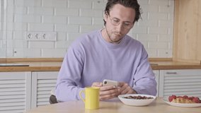 Calm handsome man in eyeglasses using smartphone while having breakfast by the table at home