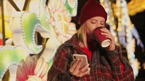 Cheerful young blonde girl wearing autumn clothes using smartphone and drinking coffee over neon lighting background