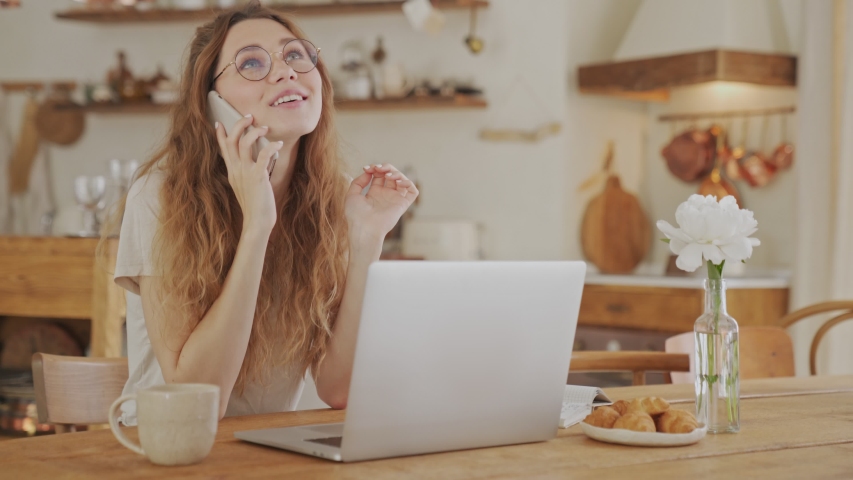 A beautiful young woman wearing glasses is talking on the phone sitting in a cozy kitchen at home