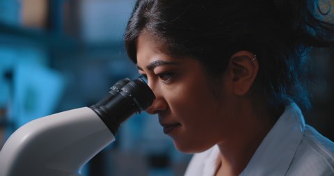 Female research scientist looking at samples under microscope.Blue lighting in a dark lab room.Close up, slider, slow motion, shot with BMPCC 4K.Biochemistry, pharmaceutical medicine, science concept
