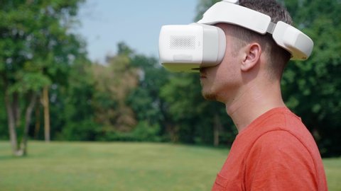 Man in virtual reality helmet turns head left and up. Guy in park in red T-shirt on background of green trees looks VR glasses. 4K footage Stock Video