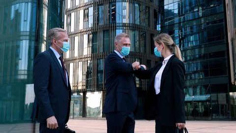 Two businessmen in medical mask meet a businesswoman also wearing a mask and use elbow bumps instead of handshake. Modern office building on the background