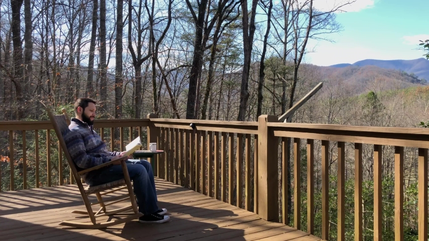 Man relaxing in a cabin in the woods while reading a book and drinking coffee in a rocking chair