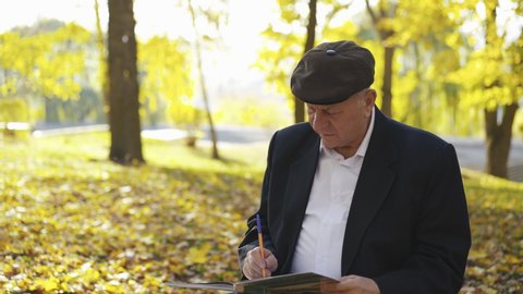 Senior man sitting on bench in an autumn park, thinking when writing and looking asides. 4K