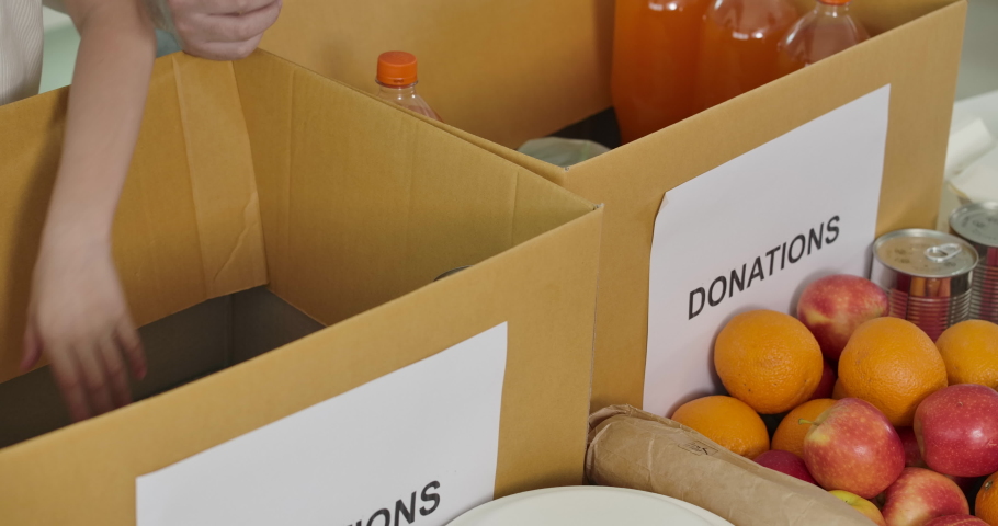 Close up shot of young food bank volunteers putting food products in donation box together as charity workers and members of the community work to the poor during the Coronavirus pandemic. Royalty-Free Stock Footage #1055587565