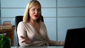 A portrait in profile of a middle-aged woman with blond hair, sitting in an office at a laptop, smiling a snow-white smile and talking on the phone on speakerphone. She has red lipstick on her lips.